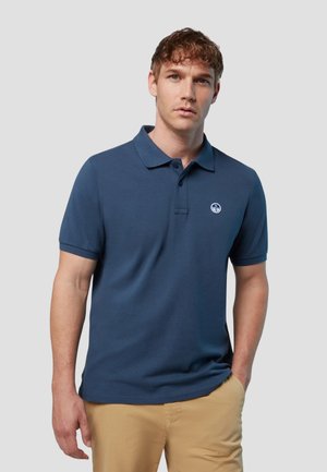 Uomo con polo a maniche corte blu navy e pantaloni beige, in piedi con una mano in tasca davanti a uno sfondo chiaro uniforme.