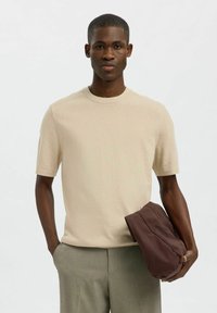 Šviesiai rudos spalvos trumpa rankovė marškinėliai, pagaminti iš lygaus audinio, su apvaliu iškirpimu. Modelis laiko rudą, minkštos tekstūros maišelį.