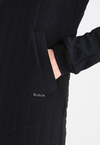 Chaqueta acolchada negra con costuras verticales, puños acanalados y un bolsillo lateral. El nombre de la marca "Bench." está bordado cerca del bolsillo.