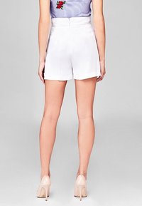 Shorts blancs ajustés avec une texture lisse, taille mi-haute et deux poches arrière. Accompagnés de chaussures à talons nus.