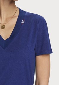 Blå T-shirt med V-hals i blødt stof med korte ærmer; har diskrete broderede detaljer nær kraven.