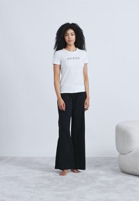 White short-sleeve t-shirt with "GUESS" logo, paired with black wide-leg pants, standing on a light grey carpet with a soft textured couch nearby.
