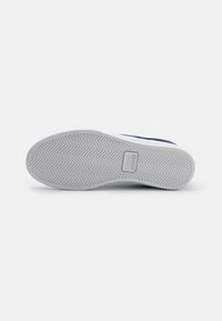 Hvite joggesko-såler med fiskebeinmønster, med en teksturert gummibunn og Lacoste-logo. Rent og enkelt design.