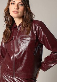 Veste en simili cuir bourgogne brillant avec un devant zippé, deux poches poitrine et un design avec col ; texture lisse et coupe cintrée.