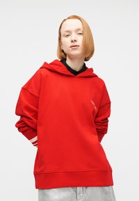 Νεαρός με κοντά κόκκινα μαλλιά που φοράει φαρδιά κόκκινη μπλούζα με το λογότυπο "Calvin Klein" και ανοιχτόχρωμο γκρι παντελόνι, μπροστά σε απλό φόντο.