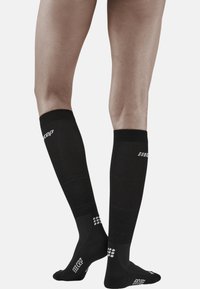 Zwarte compressiekousen met een geribbeld ontwerp, voorzien van een wit logo en patroonaccenten aan de bovenkant en de hielen. De stof lijkt glad en rekbaar.