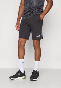 Calções atléticos pretos feitos de tecido leve, apresentando um padrão de camuflagem lateral e o logotipo branco da Puma. Combinados com sapatos pretos e meias brancas.