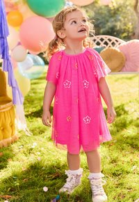 Robe rose en tissu transparent avec broderies florales. Manches courtes et design en couches. Baskets montantes scintillantes avec des accents en dentelle.