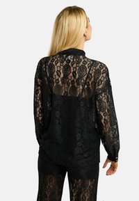 Melns mežģīņu tops ar garām piedurknēm un krekla apkakli. Rakstu veido ziedu motīvs, un aproču detaļās ir pogas, atklājot daudzslāņainu tekstūru.