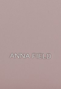 Logotipo plateado "ANNA FIELD" sobre un fondo texturizado de cuero rosa suave, centrado horizontalmente.