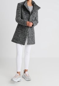 Abrigo gris texturizado con un cuello alto y dos botones, combinado con leggins blancos y zapatillas plateadas brillantes.