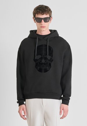 Hoodie noir avec un design de crâne texturé. Il dispose d'une capuche avec cordon de serrage, de poignets côtelés et d'une coupe décontractée. Associé à un pantalon de couleur claire.