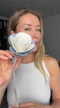 Donna con capelli lunghi e biondi tiene vicino al viso un fiore in pelle blu e bianco, indossando un top bianco senza maniche all'interno di un edificio.