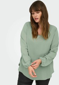 Mehka zelena bluzica z zaobljenim izrezom in zbranim rokavom. Narejena iz lahkotnega materiala z gladko teksturo in sproščenim krojem.