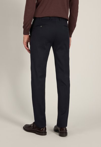 Man wearing dark navy dress pants and brown leather dress shoes standing against a plain background.