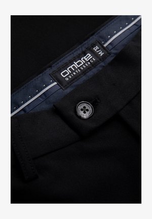 Pantalons formels noirs en tissu lisse, avec fermeture à bouton et une étiquette portant la mention "ombre quintessence". La doublure intérieure présente un motif en chevrons.