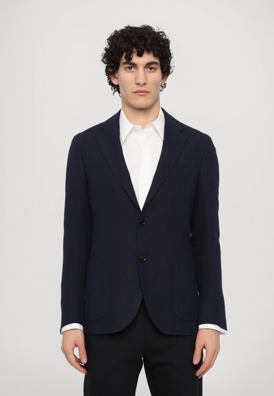 Navy blazer made of textured fabric, featuring two front pockets, two buttons, and a notched lapel, paired with a white shirt underneath.