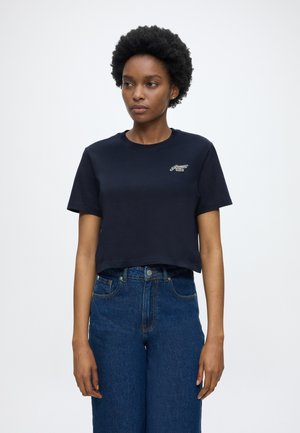 Femme aux cheveux courts et bouclés portant un t-shirt court noir avec un petit logo vert et un jean bleu taille haute, debout devant un fond blanc.