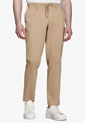 Mężczyzna w beżowych spodniach na sznurek z bocznymi kieszeniami i białych trampkach, stojący prosto na jednolitym tle.