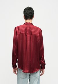 Άνδρας που φοράει ένα μακρυμάνικο, γυαλιστερό κόκκινο ριγέ πουκάμισο και ανοιχτό μπλε τζιν, στέκεται με την πλάτη στραμμένη προς την κάμερα σε απλό λευκό φόντο.