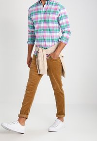 Rutig skjorta med knappar i grönt, rosa och blått med långärmar; beige manchesterbyxor; krämfärgad tröja knuten runt midjan; vita sneakers.