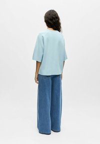 Femme aux longs cheveux bouclés portant un haut bleu clair oversize et un jean pattes d'éléphant, debout contre un fond blanc uni, de dos face au spectateur.