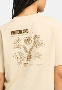 Krémové bavlněné tričko se květinovým designem v hnědé barvě na zadní straně. Text obsahuje slovo "TIMBERLAND" a názvy květin s ilustracemi.