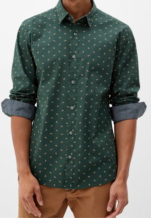 Man wearing dark green button-up shirt with small light brown patterns and rolled-up sleeves, paired with light brown pants.