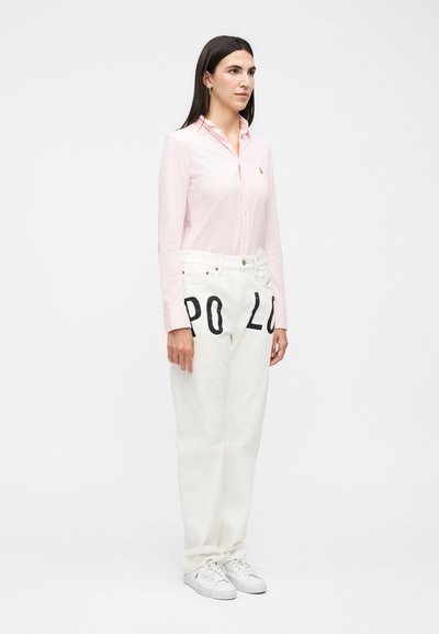 Γυναίκα που στέκεται, φορά ένα ανοιχτό ροζ πουκάμισο με κουμπιά, λευκά τζιν με μεγάλο μαύρο κείμενο "POLO" και λευκά αθλητικά παπούτσια, σε απλό φόντο.