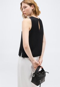 Γυναίκα με κοντά μαλλιά φορώντας μαύρο τοπ χωρίς μανίκια και λευκό παντελόνι, κρατώντας μια μικρή μαύρη τσάντα χειρός πίσω από την πλάτη της.