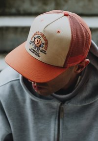 Vrachtwagenpet met een beige voorkant en oranje klep; voorzien van een geborduurde perzikkleurige afbeelding en de tekst "PEACH PERFECT." Gaasachterkant voor ventilatie.