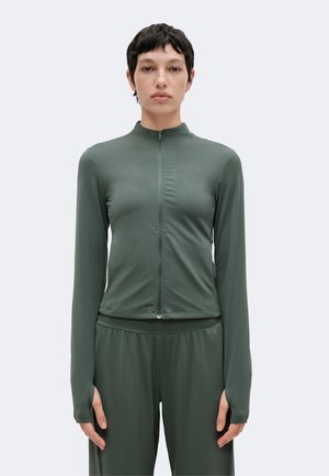 Femme aux cheveux courts et foncés, portant un haut vert ajusté à manches longues avec fermeture éclair et un pantalon ample assorti, debout devant un fond clair.