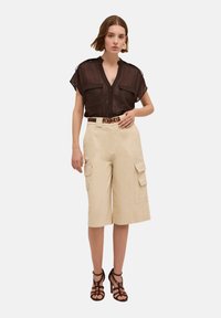 Schwarzes, durchsichtiges kurzärmliges Hemd mit zwei Brusttaschen, beige Cargo-Hose mit Seitentaschen und schwarze, Riemchen-High Heels. Brauner Gürtel als Akzent.