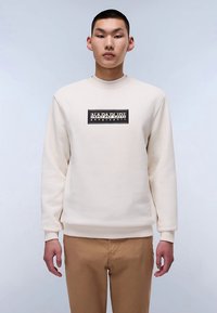 Béžová mikina s okrúhlym výstrihom a rebrovanými manžetami, na prednej strane čierne obdĺžnikové logo. Nosí sa s svetlohnedými nohavicami.