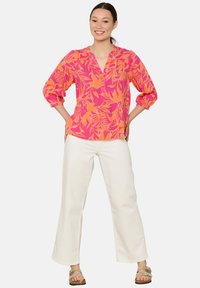 Rosa und orangeblumiges Oberteil mit V-Ausschnitt und Puffärmeln, getragen mit weißen, weiten Hosen und beigen Sandalen vor einem schlichten Hintergrund.