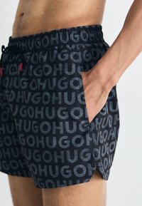 Shorts de baie negre cu un model repetat de logo "HUGO" în albastru, cu talie elastică și buzunare laterale.