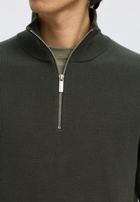 Mand iført olivengrøn vaffelstrikket sweater med sølv halv lynlås og ribbet høj krave over en lysere grøn t-shirt med rund hals.