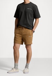 Czarna koszulka z okrągłym dekoltem i brązowe bawełniane spodenki ze sznurkiem. Białe logo na lewej piersi i na spodenkach. Model ma na sobie białe trampki i szare skarpetki.