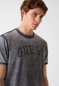 Szara koszulka wykonana z miękkiej, teksturowanej tkaniny z dużym, wyblakłym czarnym logo „GUESS” na przodzie oraz krótkimi rękawami.