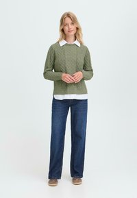 Olijfgroen gebreide trui met een gestructureerd patroon, gecombineerd met een witte blouse met kraag, blauwe jeans en bruine schoenen.