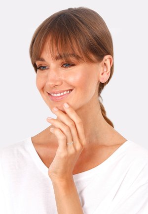 Silver ring on a hand with light nail polish, worn by a woman in a white shirt. She has brown hair and a subtle smile.