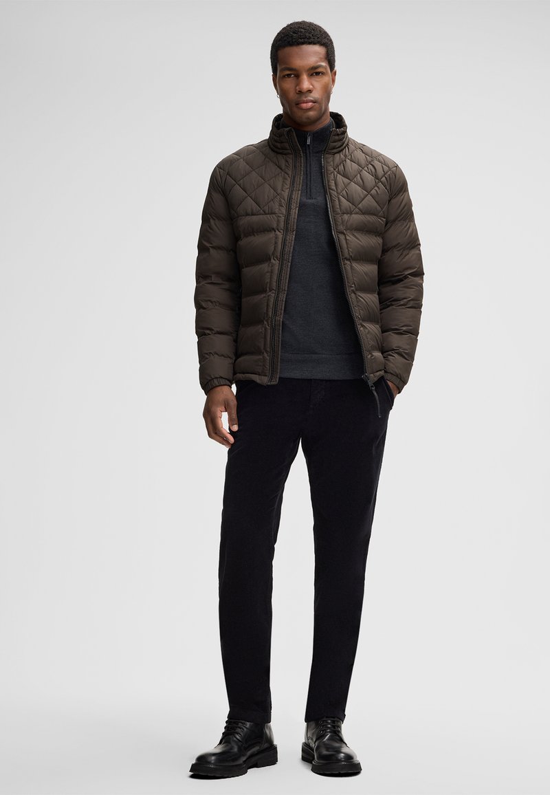 Giacca trapuntata marrone imbottita con collo alto, indossata sopra un maglione scuro. Abbinata a pantaloni neri e stivali neri stringati.