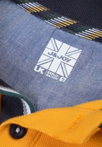 Kraagpoloshirt met gele textuurstof, marineblauwe accenten en een label met een Union Jack-ontwerp, wat aangeeft dat het merk J&JOY is.