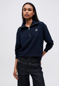 Femeie îmbrăcată într-un hanorac navy blue cu fermoar la jumătate, având un logo alb pe piept și blugi de culoare albastru închis, stând în fața unui fundal deschis simplu.