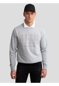 Mand iført sort kasket, hvid skjorte med krave under grå sweatshirt med teksten "BIG STAR" og sorte bukser, stående foran en ensfarvet baggrund.