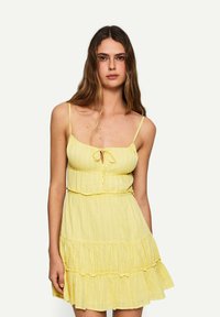 Abito giallo senza maniche con dettaglio a nodo sul davanti, caratterizzato da una vita arricciata e un orlo a strati con ruches. Tessuto leggero con una sottile texture.