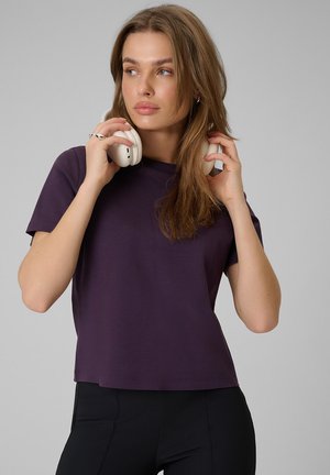 Młoda kobieta z długimi włosami, ubrana w ciemnofioletowy t-shirt, trzyma białe słuchawki nausznikowe blisko szyi, patrząc na bok.