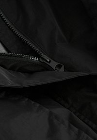 Svart lättviktig jacka med synlig dragkedja. Materialet verkar jämnt och något texturerat, med en subtil lyster och rena sömmar.