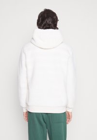 Vit fleece hoodie med hög krage, texturerat tyg och ribbad mudd, tillsammans med gröna byxor. Bakifrån, inga synliga varumärken.