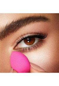 Close-up van een oog met een bruine iris, lange wimpers en een zachtroze make-up spons die tegen de huid wordt gehouden. Glad, licht huidtype.